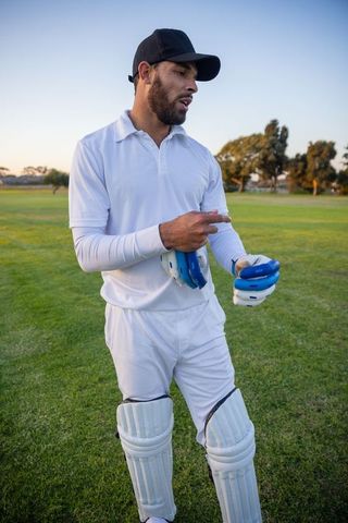 Cricketer standing on sunlit outfield wearing white kit and pads holding blue gloves