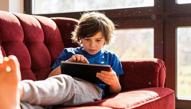 Young boy lounging on red sofa using tablet in sunlit room with dust motes