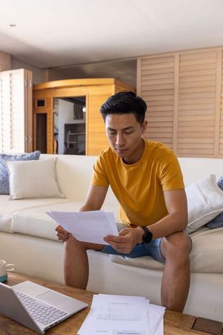 Asian young man reading bills and working on laptop on couch minimalist sunlit workspace