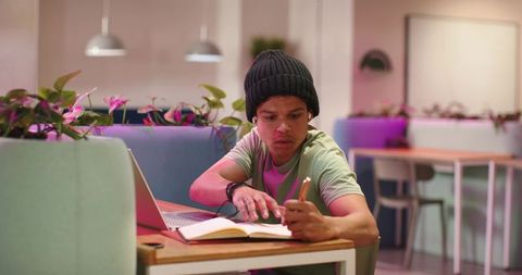 Young man studying and writing in modern coworking cafe with laptop and plants