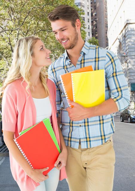 Two teenage students standing outdoors in an urban environment, holding colorful books and smiling at each other. Ideal for use in educational materials, advertisements for school supplies, or promotional content for academic institutions. The image conveys themes of friendship, learning, and youthful energy.