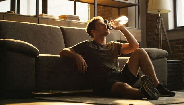 Sunlit post-workout man drinking water while resting on loft floor beside gray sofa and brick wall