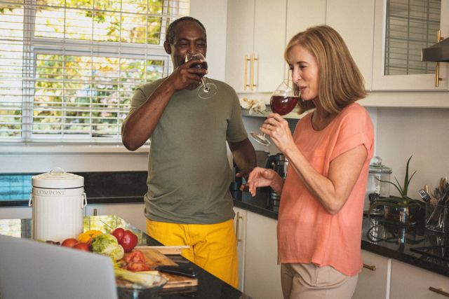Senior couple enjoying red wine in a modern kitchen, creating a relaxed and intimate atmosphere. Ideal for use in articles or advertisements about senior living, home life, quarantine activities, or promoting a healthy and happy lifestyle for older adults.