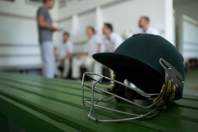Cricket helmet resting on green bench in locker room with blurred team members in background. Ideal for sports-related articles, cricket gear advertisements, team preparation visuals, and athletic training content.
