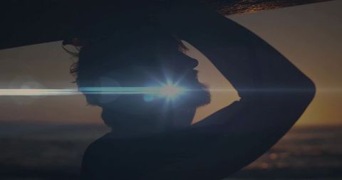 Silhouetted man carrying surfboard at sunset on coastline with dramatic lens flare