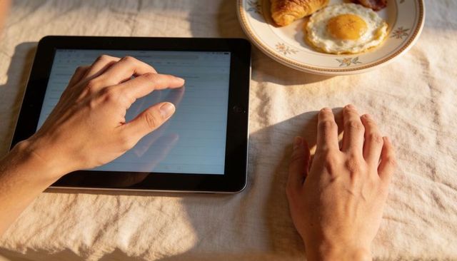 Morning tablet browsing with hands tapping touchscreen beside egg and croissant