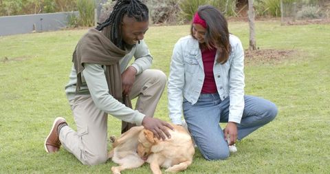 Multicultural couple kneeling on lawn petting dog, casual outdoor affection and bonding