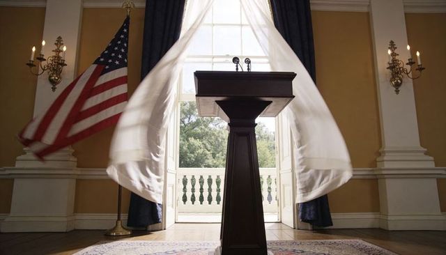 Dramatic dark wood podium framed by billowing sheer curtains, twin microphones and american flag
