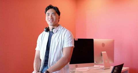Young asian man leaning on desk in pink modern workspace with dual desktops and headphones