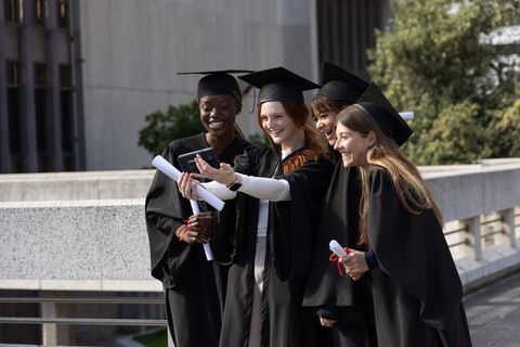 Female graduates taking selfie on campus terrace celebrating diplomas smiling together