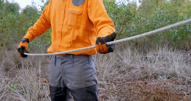 Forest worker holding rope in outdoor environment. Person in safety gloves and orange jacket performing manual labor in forest setting. Ideal for concepts related to forestry work, outdoor labor, safety gear, and environmental conservation.