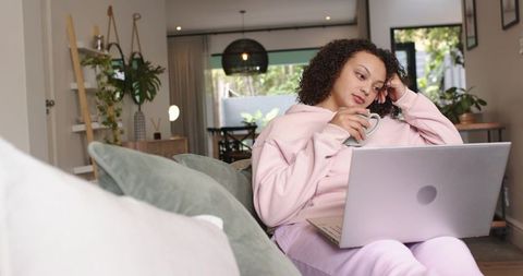 Young woman relaxing on sofa with laptop and mug, working from home in cozy living room