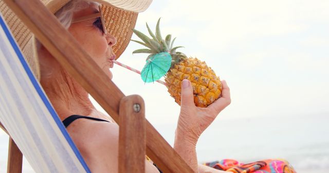 Senior woman enjoying a tropical vacation, sipping a pineapple cocktail with a straw while relaxing on a beach chair. This can be used for promoting vacation destinations, retirement lifestyle, travel brochures, summer activities, or health and wellness blogs targeting active seniors.