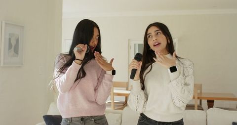 Two diverse female friends singing karaoke in cozy living room