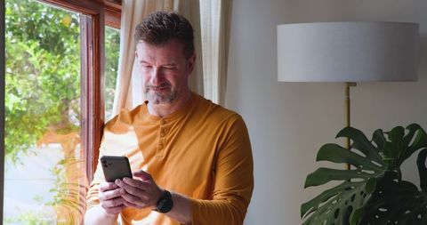Middle-Aged Man in Mustard Henley Engaged with Smartphone Indoors