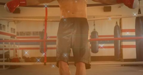 Resting shirtless boxer leaning on ropes wearing red gloves and dark shorts in boxing ring