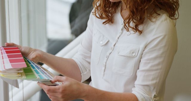 Redhead woman wearing white shirt holding a variety of color swatches. Suitable for content related to interior design, home renovation, creative profession marketing, and documenting the process of selecting colors for decoration or remodeling projects.