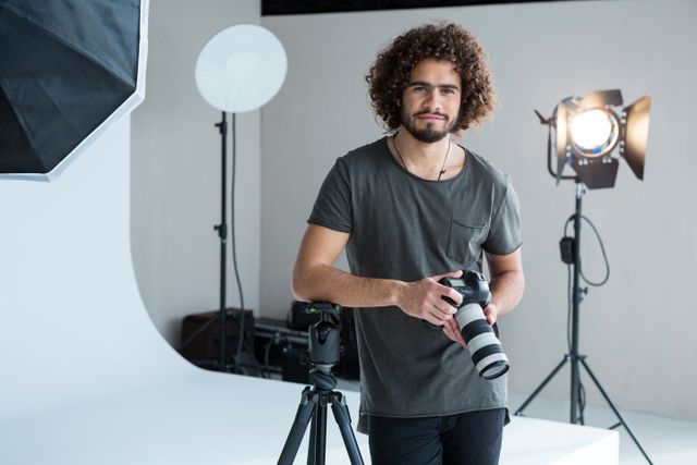 Young male photographer with curly hair standing in a professional studio holding a camera. He is smiling and surrounded by lighting equipment and a tripod. Ideal for use in articles about photography, creative professions, or studio setups.