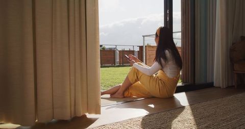 Asian woman sitting on sliding door threshold holding smartphone gazing at sunlit backyard