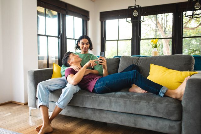Multiracial young man showing smartphone to boyfriend while lying on his lap on sofa in living room. Technology, relaxing, unaltered, love, togetherness, homosexual, lifestyle and home concept.