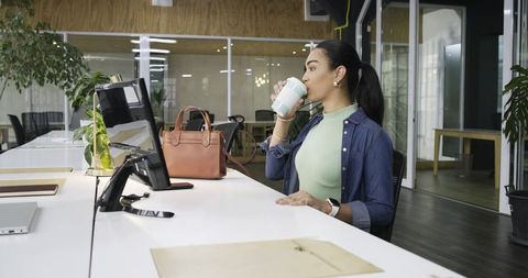 Indian woman sipping travel mug at modern coworking desk with handbag and monitor