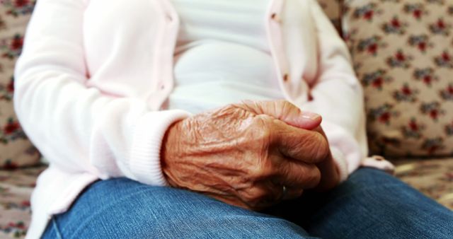 Elderly woman with wrinkled hands seen sitting on a couch in a living room, wearing a light pink cardigan and jeans. Perfect for use in articles or promotions related to aging, senior lifestyle, elder care, or healthcare services. Can also be used in visual content about family and home care.