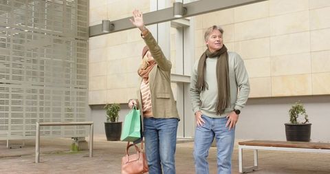 Mature couple waving, holding shopping bags, standing in urban plaza, casual autumn fashion