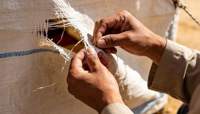 Repairing torn tarpaulin with calloused hands twisting twine under sunlit khaki sleeves