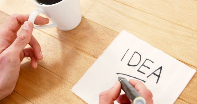 Person writing 'IDEA' in bold letters on a napkin while holding a cup of coffee in the background. Perfect for illustrating creative processes, brainstorming sessions, business concepts, casual work environments, and inspirational content.