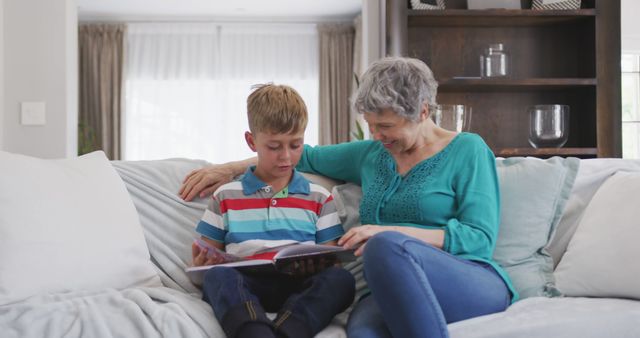 Elderly woman bonding with young boy while reading storybook together on couch in cozy living room. Ideal for articles on family bonding, grandparent-grandchild relationships, or learning and literacy activities at home.