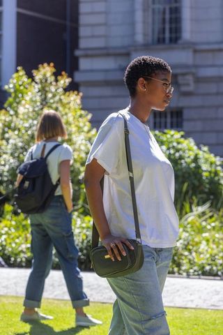 African woman walking campus wearing white tee, glasses and crossbody bag in sunlit courtyard