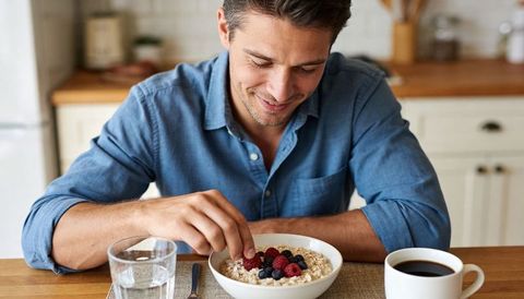Man adding berries to oatmeal at kitchen table with coffee