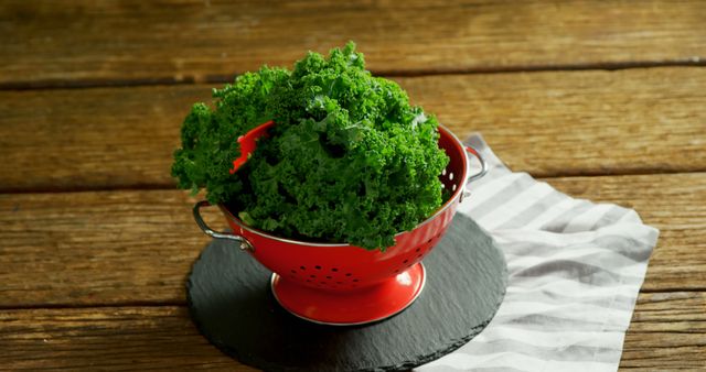 A vibrant bunch of fresh kale sits in a red colander on a rustic wooden table, with copy space. Kale is known for its health benefits and is a popular ingredient in nutritious recipes.