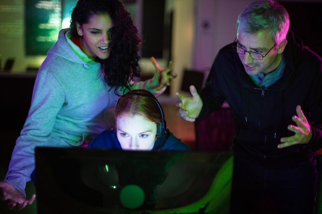 This image captures a team of coworkers collaborating late at night in a creative office environment. The focus is on a woman wearing headphones, typing on a computer keyboard, while two colleagues stand by her, actively brainstorming. Ideal for illustrating concepts of teamwork, late-night productivity, creative problem solving, and collaborative work in tech or creative industries.
