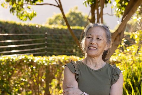 Senior asian woman smiling in sunlit garden wearing olive top and fitness wristband