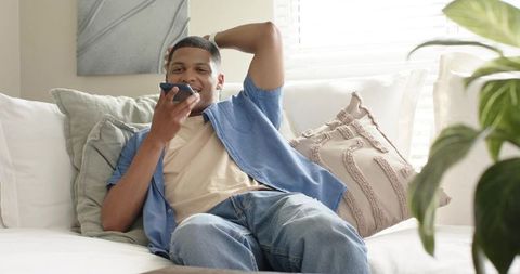 African american man reclining on sofa speaking into smartphone in cozy pastel living room