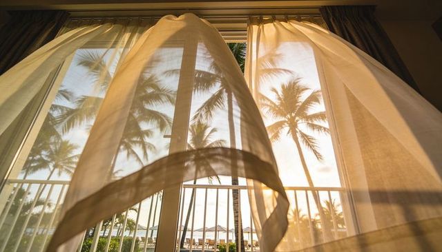 Billowing sheer curtain framing tropical palm-tree balcony at golden sunrise resort