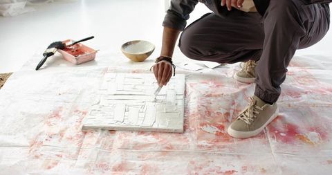 Indian artist spreading white texture paste with palette knife over red-splattered drop cloth