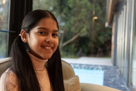 Indian girl smiling in sunlit sunroom sitting by pool wearing pink turtleneck and pigtails