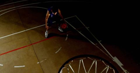 Overhead dramatic basketball player dribbling under spotlight on indoor court with hoop