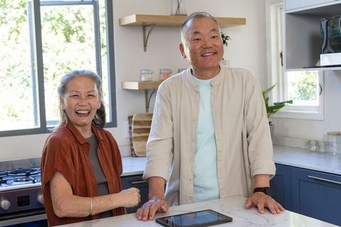 Senior asian couple smiling and standing at marble countertop using tablet in sunlit kitchen