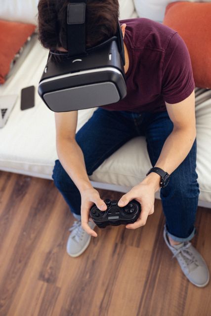 Teenage boy wearing a virtual reality headset and holding a game controller while sitting on a couch at home. Ideal for use in articles or advertisements related to modern technology, gaming, virtual reality experiences, and youth entertainment. Can also be used to illustrate concepts of immersive technology and home leisure activities.