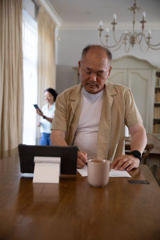 Senior asian man signing documents at table while partner using smartphone in living room