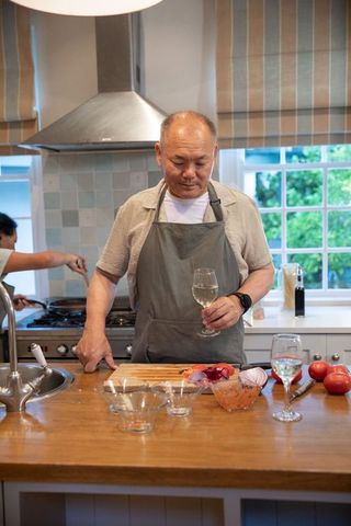 Mature chinese man preparing evening dinner in kitchen holding wine glass and chopping veg