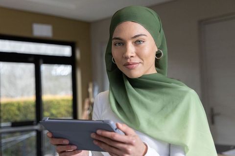 Confident young woman wearing green hijab holding tablet in modern home workspace
