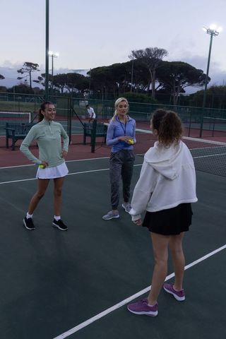 Female players practicing tennis at dusk on outdoor court with coach and floodlights