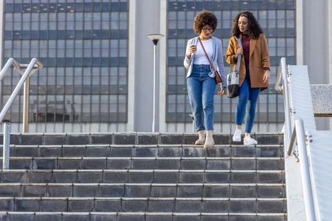 Two diverse women walking down urban steps carrying coffee and bags in business-casual