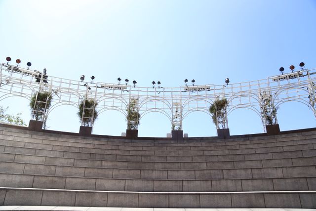 Wide shot of an outdoor amphitheater with tiered seating and minimalistic metal arch structure overhead. Scene is illuminated by sunlight under a clear blue sky. Ideal for use in articles about architectural design, outdoor venues, public performance spaces, or travel destinations.