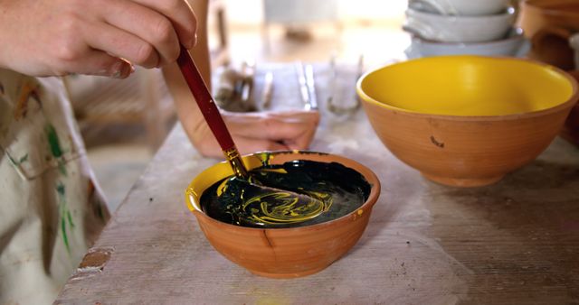 Artist's hand mixing yellow and black paint in a clay pottery bowl using a brush. Useful for themes on creativity, art projects, handicrafts, artisan workshops, painting techniques, and studio work. Could be used in content about DIY projects, instructional art materials, or promoting art classes and studios.