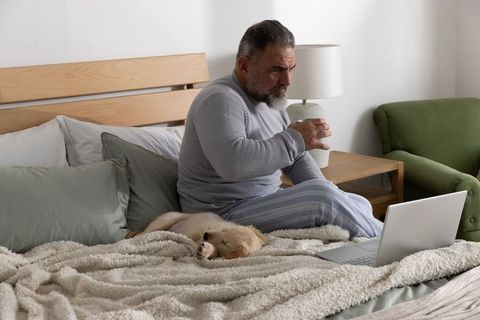 Senior man in pajamas holding mug and using laptop on cozy bed with sleeping golden puppy